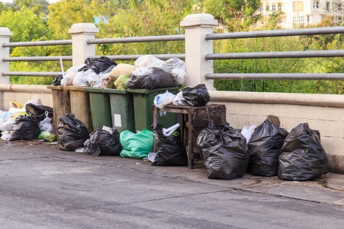 A collection of black and green rubbish bags, along with a few white and transparent bags, are placed on and around a concrete pavement beside a metal railing, with some bags leaning against a green wheelie bin. The area appears to be an urban street scene, possibly near a residential or commercial property in West Wickham, with trees and greenery visible in the background. The bags are filled with waste materials, and the scene indicates a waste removal activity typical for municipal or commercial rubbish collection services, with Commercial Waste West Wickham managing waste disposal in the area. The pavement is dark asphalt, and the bags are various sizes and shapes, some tied tightly, others loosely filled, providing a realistic view of rubbish awaiting collection from a local waste management service location.