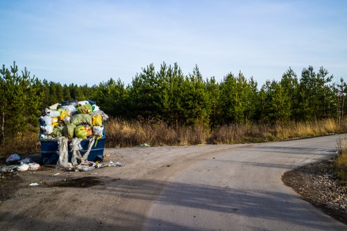 A large blue waste skip positioned on the side of a rural dirt road, filled with a variety of discarded rubbish including yellow and white plastic bags, crumpled cardboard boxes, and miscellaneous debris, with additional loose waste scattered on the ground nearby. The skip is situated in an open area with sparse vegetation and a line of dense green trees in the background under a partly cloudy sky, indicating a natural environment outside of an urban setting. The scene reflects a typical site for rubbish collection or disposal in the West Wickham area, consistent with services provided by Commercial Waste West Wickham, highlighting the importance of professional waste removal in managing refuse in rural and suburban landscapes.