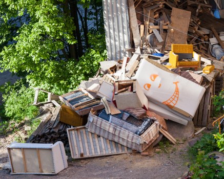 An outdoor scene showing a pile of discarded furniture and waste materials, including a striped fabric-covered sofa, wooden chairs, and a cardboard box with orange printed design, all situated on a paved area next to a corrugated metal wall. The furniture appears weathered and is overlapping, with some items leaning against the wall. To the left, there are green leafy trees providing natural background contrast, and the environment suggests a typical waste removal scenario possibly in West Wickham. The image emphasizes the type of bulky household rubbish that Commercial Waste West Wickham would collect and dispose of. The scene is illuminated by natural daylight, highlighting the textures and colors of the various objects, with the surrounding greenery and wall framing the clutter in the foreground. This visual captures the nature of furniture clearance and rubbish removal services in a residential or commercial setting within the local area.