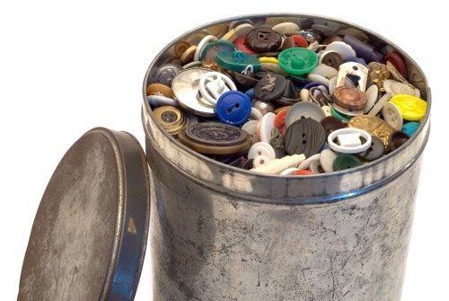 A round metal container filled with a large collection of plastic buttons of various sizes, shapes, and colors, including shades of blue, white, black, red, yellow, and green. The container, which has a slightly tarnished and weathered appearance, is situated outdoors on a paved surface, with its matching metal lid leaning against it on the left side. The background features a blurred outdoor environment, suggesting it is part of a waste collection or disposal setup, possibly at a commercial waste removal site in West Wickham. The lighting appears natural, highlighting the diverse textures and vivid colors of the buttons within the container, which are contributing to the overall scene involving rubbish collection and waste management services provided by Commercial Waste West Wickham.