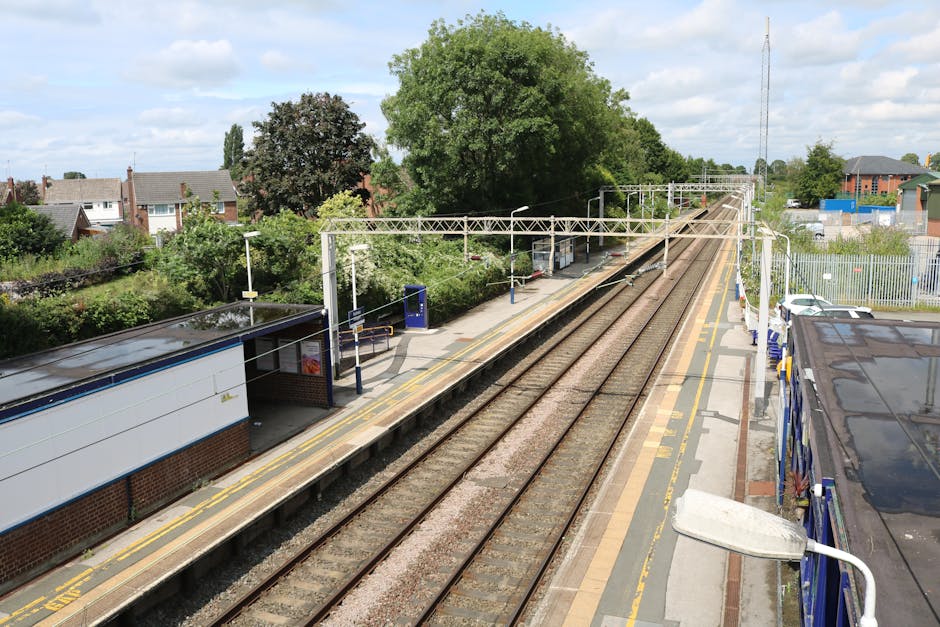 An elevated view of a railway station platform with a double track running through the centre, flanked by a small waiting shelter on the left with a dark, flat roof and brick base, and a modern street