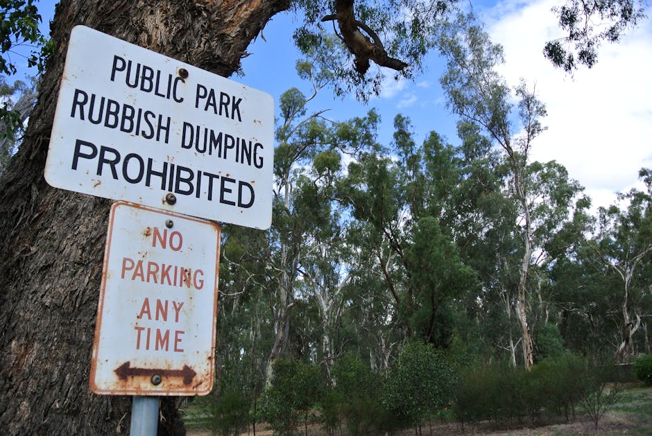 A rectangular white metal sign with black text mounted on a red brick wall. The sign reads 'NO DUMPING OF RUBBISH' with the word 'DUMPING' partially obscured or misaligned. The brick wall features a t