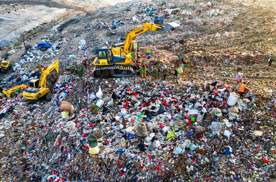 A large landfill site with multiple yellow excavators sorting and managing waste on a sloped, uneven terrain covered in various types of trash, including plastic, paper, and organic materials, with a 